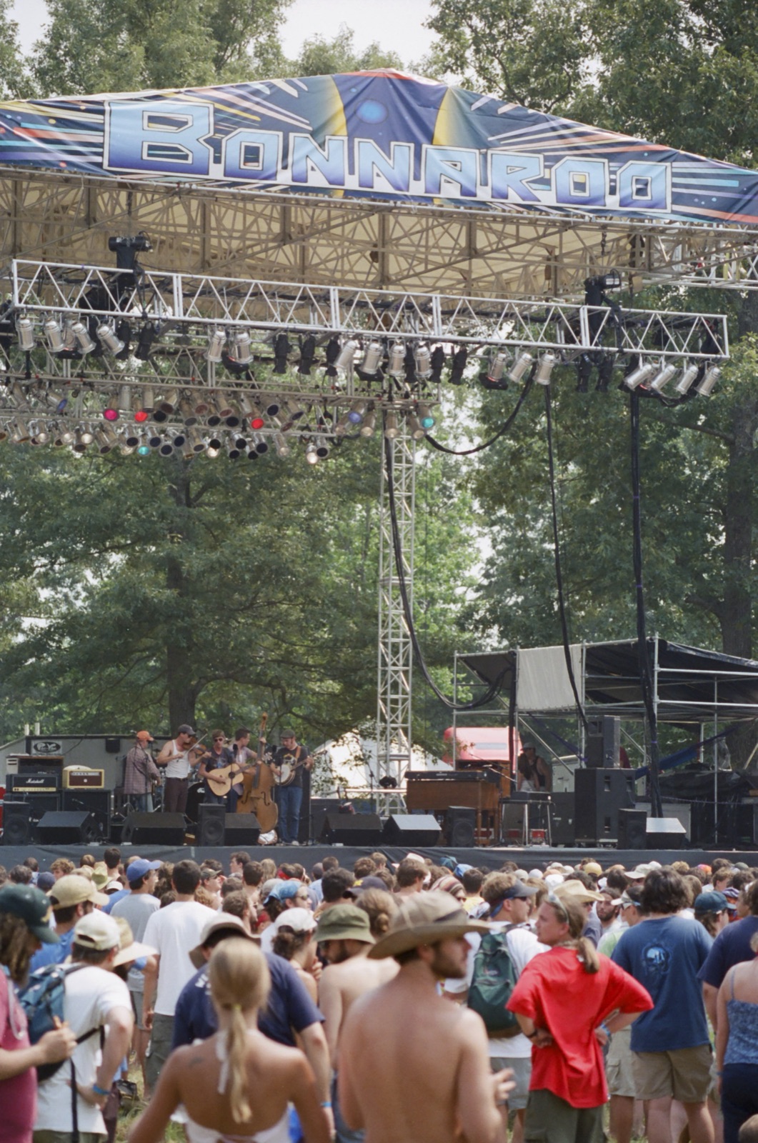 A string band plays under a big Bonnaroo banner and lighting rig while a sunburned crowd fills the field in front.